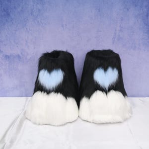 May include: Pair of black and white faux fur boots with a light blue heart design. The boots have a fluffy texture and are designed to resemble animal paws. The boots are a unique accessory for cosplay or costume events.