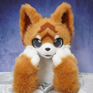 May include: A plush animal head mask featuring orange and white faux fur with large, dark, glossy eyes. The design includes pointed ears, a white muzzle, and brown markings on the forehead. The mask is crafted with soft, textured material.