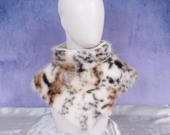 Puede incluir: Un cuello de piel sintética con cuello alto. El cuello presenta un patrón de manchas blancas, marrones y negras, similar a un leopardo de las nieves. Se muestra sobre una cabeza de maniquí blanca sobre un fondo azul.