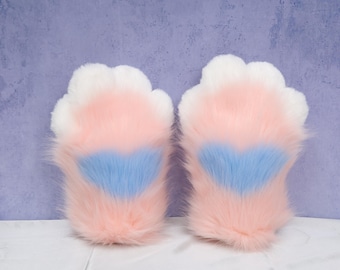 Puede incluir: Un par de guantes de pata de felpa en rosa, blanco y azul. Los guantes tienen una textura esponjosa con un diseño en forma de corazón en el centro de las almohadillas de la pata. El fondo es de color púrpura claro.