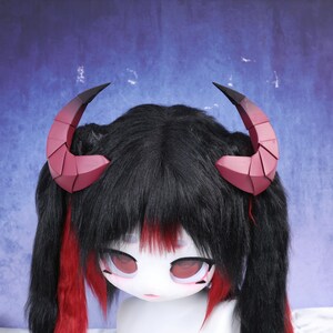 May include: A doll with black hair, red highlights, and large red eyes. The doll is wearing a pair of burgundy and black geometric horns. The doll's face is white with red accents, and it has a neutral expression.