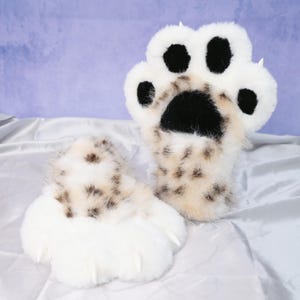 May include: A pair of plush paw mittens, featuring a white and tan spotted design. The mittens have black pads and white claws, resembling a large animal paw. The soft, furry texture is visible.