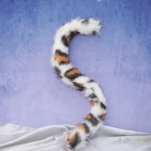 May include: A plush cat tail in white, black, and orange spotted pattern. The tail is curved and fluffy, with a soft texture. The tail is likely a costume accessory.