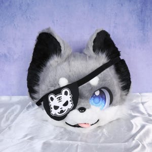 May include: A plush, furry wolf head with gray and black fur, large blue eyes, and a black eye patch featuring a white tiger design. The tongue is pink, and the ears are upright.