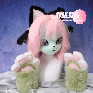 May include: A furry cat-like mask with pink hair and black ears. The mask has large, expressive eyes and a green face. Matching green and white paw gloves are in front of the mask. The text "ONLY ONE AVAILABLE EXCLUSIVE PIECE" is on a pink cloud.