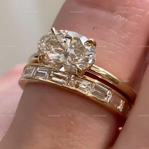 May include: Close-up of a gold ring set featuring a large oval-cut diamond and a band with baguette-cut diamonds. The ring is set on a gold band. The diamonds sparkle in the light, showcasing the ring's intricate design.