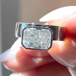 May include: A silver ring featuring a large, rectangular, clear gemstone with a black bezel setting. The ring band is wide and polished, reflecting light. The gemstone sparkles, showcasing its facets.