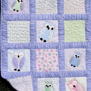 May include: A purple, blue, and green baby quilt with a patchwork design featuring owls. The quilt has a ruffled border and is made of soft, quilted fabric.