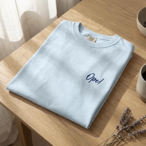 May include: A light blue t-shirt with the word "Ope!" embroidered in navy blue script. The shirt is folded neatly on a wooden surface. A small tag is visible near the collar. The shirt is part of a clothing collection.