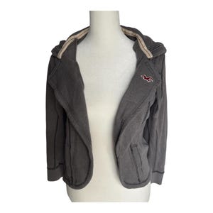 May include: A gray zip-up jacket with a hood and two front pockets. The jacket has a red logo on the chest and a contrasting beige lining around the hood and collar. The jacket is displayed on a white mannequin.