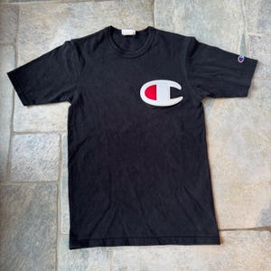 Op de afbeelding: Zwart Champion T-shirt met een groot wit, rood en grijs logo op de borst. Het shirt heeft korte mouwen en een ronde hals. Een kleiner Champion-logo staat op de linkermouw. Het shirt is gemaakt van een zacht, comfortabel materiaal.