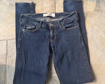 May include: Dark blue denim jeans with a button and zipper closure. The jeans have a classic five-pocket design, a small embroidered logo on the front pocket, and a slim fit. The jeans are folded on a stone surface.