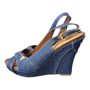 May include: Blue denim wedge sandals with a peep toe design. The sandals feature a slingback strap and a gold-tone buckle. The wedge heel is approximately 4 inches high. The Guess logo is visible on the front.