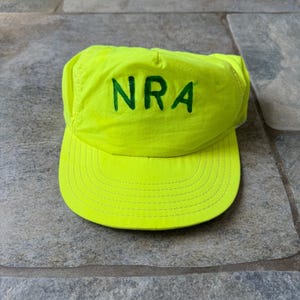 Puede incluir: Gorra de béisbol de color amarillo brillante con las letras "NRA" en verde. La gorra tiene una visera curva y está hecha de un material ligero. El sombrero está sobre una superficie de piedra.