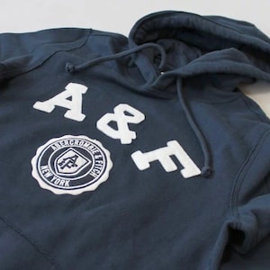 May include: Navy blue hoodie featuring white embroidered 'A & F' lettering and a circular patch reading 'Abercrombie & Fitch New York'. The hoodie has a drawstring hood.