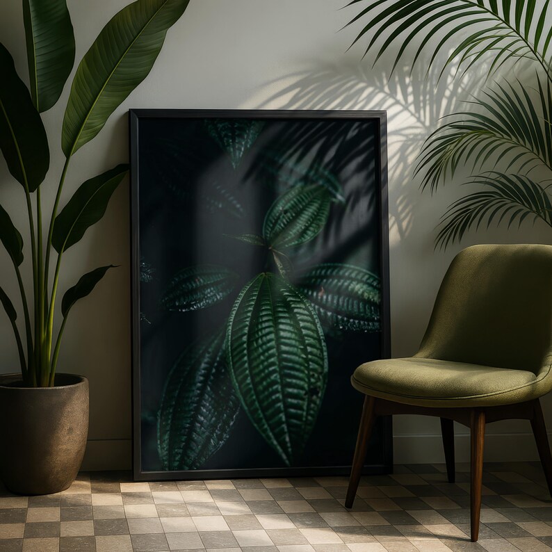 May include: A framed botanical print featuring large, textured green leaves. The artwork is set against a dark background, creating a dramatic contrast. A green chair and potted plant are in the foreground, adding to the natural theme.