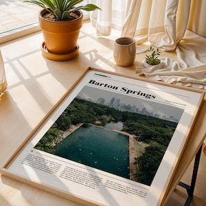 May include: A framed print titled "Barton Springs" featuring a photograph of a natural swimming pool surrounded by trees, with a city skyline in the background. The print is displayed on a wooden surface near a window.
