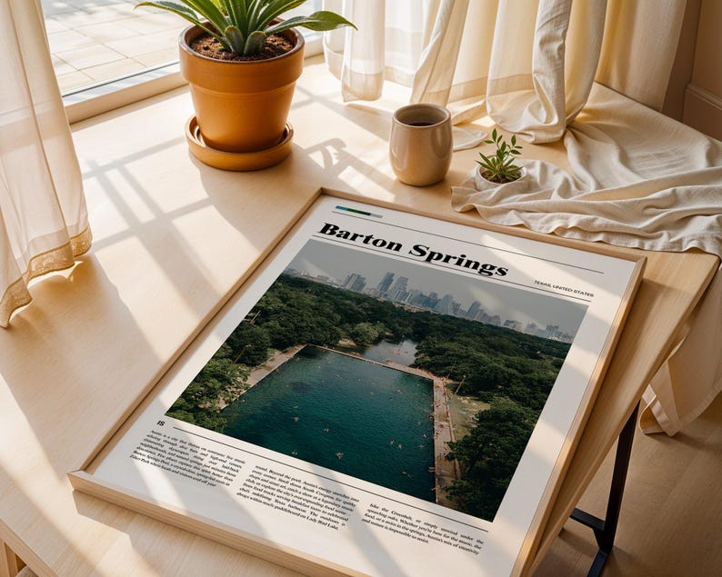 May include: A framed print titled "Barton Springs" featuring a photograph of a natural swimming pool surrounded by trees, with a city skyline in the background. The print is displayed on a wooden surface near a window.