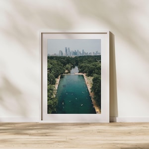 May include: Framed aerial photograph of a large, rectangular swimming pool filled with turquoise water, surrounded by lush green trees. A city skyline is visible in the distance under a cloudy sky. The photograph is displayed in a white frame.