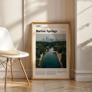 May include: A framed print with the text "Barton Springs" at the top. The image shows a pool surrounded by trees with a city skyline in the background. The frame is gold, and the print is displayed next to a white chair.