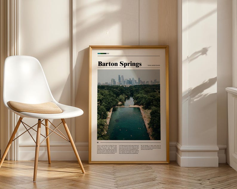 May include: A framed print with the text "Barton Springs" at the top. The image shows a pool surrounded by trees with a city skyline in the background. The frame is gold, and the print is displayed next to a white chair.