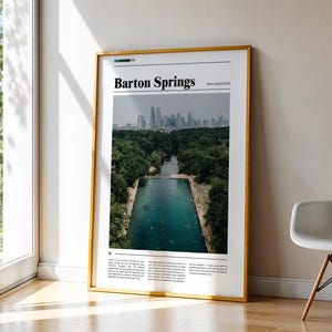 May include: A framed poster with a gold border, leaning against a wall. The poster features a photograph of Barton Springs in Austin, Texas, with the text "Barton Springs" at the top. The image shows a pool surrounded by trees and a city skyline in the background.