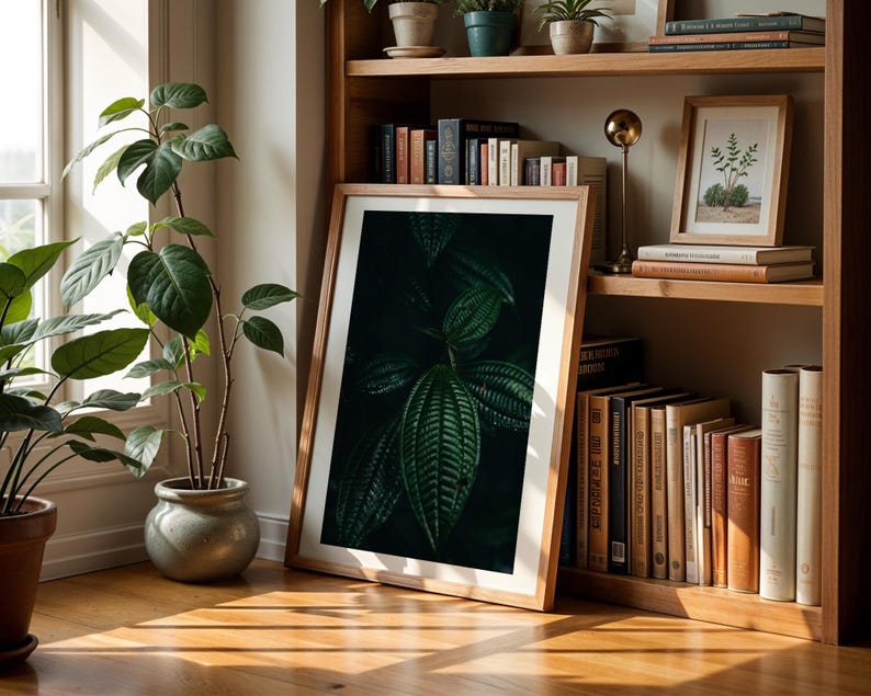 May include: A framed botanical print featuring a close-up of dark green leaves, leaning against a wooden bookshelf. The room is illuminated by natural light, with potted plants and books adding to the decor. The frame is a light brown color.