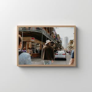 Könnte beinhalten: Ein gerahmtes Foto einer Straßenszene mit Menschen, die gehen. Eine Person in einer braunen Jacke und einem weißen Hut geht von der Kamera weg. Gebäude mit Balkonen und Flaggen säumen die Straße. Ein Auto ist im Hintergrund.