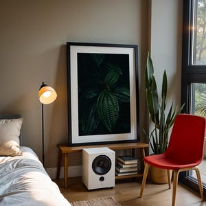 May include: A modern interior scene featuring a framed botanical print, a white speaker, and a red chair. A floor lamp with a yellow lampshade illuminates a bed with a beige pillow. A tall green plant and a window with natural light complete the room.