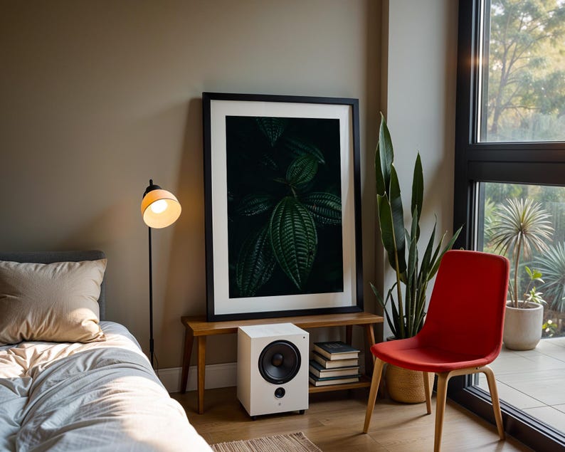 May include: A modern interior scene featuring a framed botanical print, a white speaker, and a red chair. A floor lamp with a yellow lampshade illuminates a bed with a beige pillow. A tall green plant and a window with natural light complete the room.