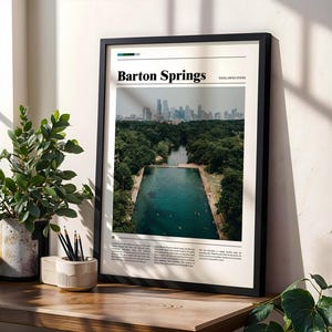 May include: A framed print featuring a photograph of Barton Springs, with the text "Barton Springs" at the top. The image shows a natural swimming pool surrounded by trees, with a city skyline in the background. The frame is black.