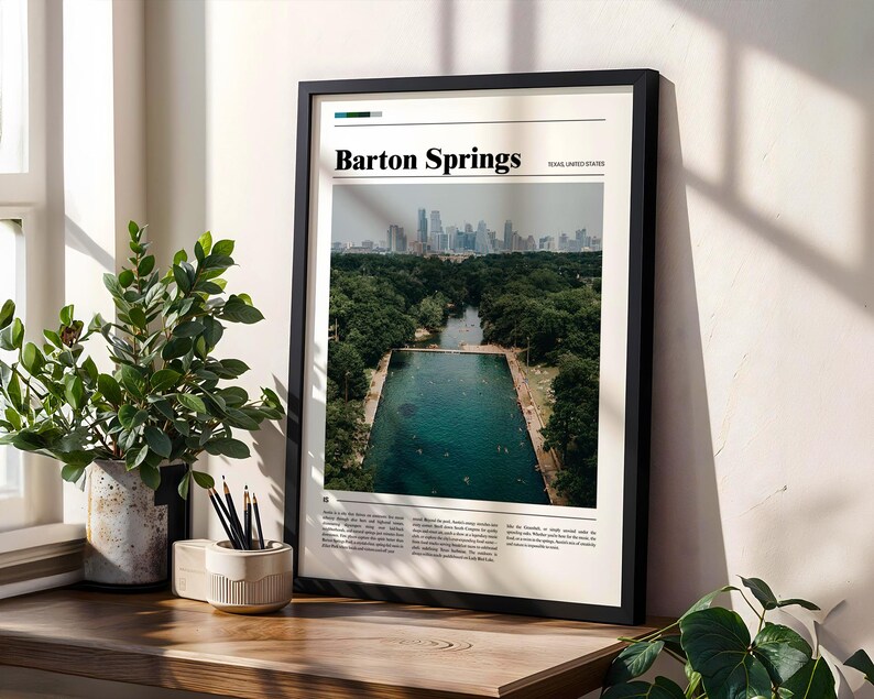 May include: A framed print featuring a photograph of Barton Springs, with the text "Barton Springs" at the top. The image shows a natural swimming pool surrounded by trees, with a city skyline in the background. The frame is black.