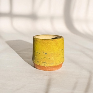 May include: A small ceramic cup with a cylindrical shape, featuring a vibrant yellow glazed upper section with subtle dark speckles and a contrasting unglazed terracotta-colored base. The interior of the cup is a smooth, light cream color.