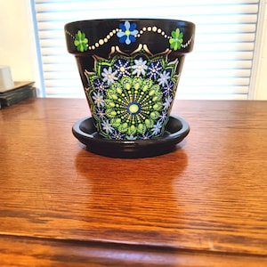 May include: A black ceramic flower pot with a matching saucer. The pot is decorated with a hand-painted mandala design in shades of blue, green, and white. The rim features small flowers and white dots. A decorative item for home decor.