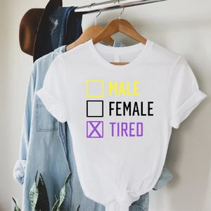 May include: White t-shirt with the text "MALE", "FEMALE", and "TIRED" in yellow, black, and purple. The word "TIRED" has a purple "X" in the check box. The shirt is hanging on a wooden hanger with other clothing in the background.
