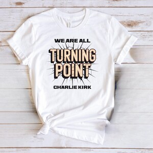 A white t-shirt with a graphic design featuring the text "WE ARE ALL TURNING POINT CHARLIE KIRK" printed on it.
