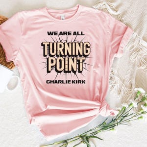 A pink t-shirt with the text "WE ARE ALL TURNING POINT CHARLIE KIRK" printed on it, along with a coffee mug and some flowers on a bed.