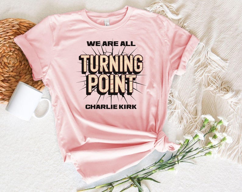 A pink t-shirt with the text "WE ARE ALL TURNING POINT CHARLIE KIRK" printed on it, along with a coffee mug and some flowers on a bed.