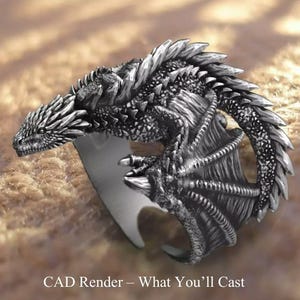 May include: A silver dragon ring with intricate detailing. The dragon wraps around the finger, featuring textured scales, wings, and a detailed head. The ring is set against a neutral background. Text at the bottom reads "CAD Render - What You'll Cast."