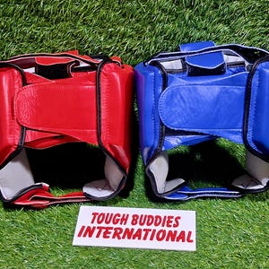 Op de afbeelding: Twee bokshoofdbeschermers, &eacute;&eacute;n rood en &eacute;&eacute;n blauw, worden getoond op een groene ondergrond. De hoofdbeschermers hebben een gewatteerd ontwerp met verstelbare bandjes. De tekst "TOUGH BUDDIES INTERNATIONAL" staat op een witte rechthoek.