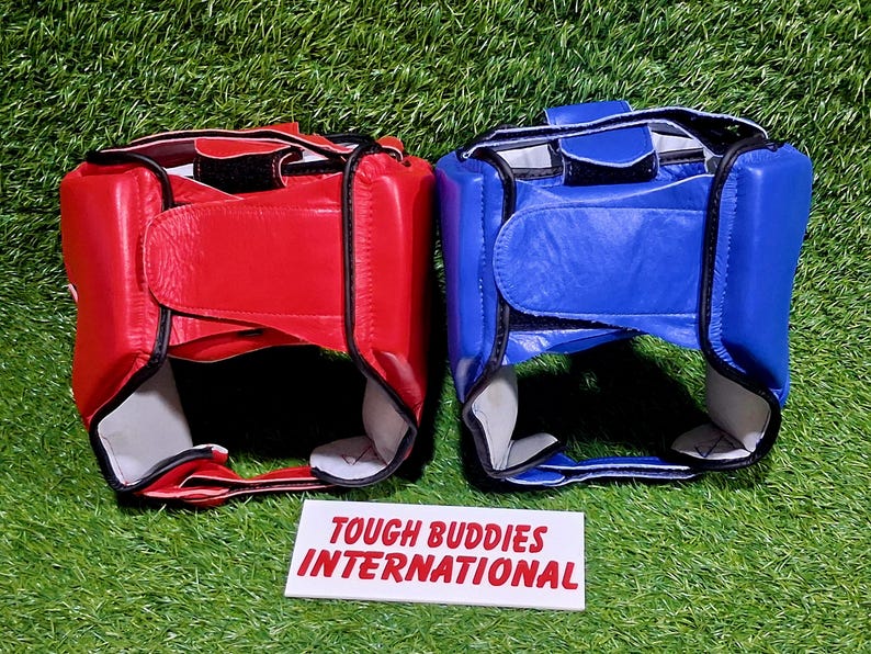 Op de afbeelding: Twee bokshoofdbeschermers, &eacute;&eacute;n rood en &eacute;&eacute;n blauw, worden getoond op een groene ondergrond. De hoofdbeschermers hebben een gewatteerd ontwerp met verstelbare bandjes. De tekst "TOUGH BUDDIES INTERNATIONAL" staat op een witte rechthoek.