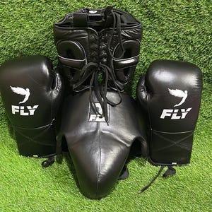 K&ouml;nnte beinhalten: Schwarzer Boxkopfschutz, ein Tiefschutz und zwei Boxhandschuhe liegen auf einer gr&uuml;nen Oberfl&auml;che. Die Handschuhe und der Kopfschutz haben wei&szlig;e Akzente und das Wort "FLY" mit einem Fl&uuml;gelmotiv. Der Kopfschutz hat einen Schn&uuml;rverschluss.