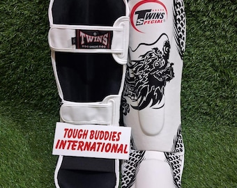 Könnte beinhalten: Paar weiße und schwarze Schienbeinschützer mit dem Logo "Twins Special" und einem schwarzen Drachendesign. Die Schienbeinschützer haben weiße Riemen und den Text "Tough Buddies International".