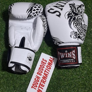 May include: White boxing gloves with black dragon and feather designs. The gloves have the word "TWINS" printed on them, along with the text "16 oz". A white card with the text "TOUGH BUDDIES INTERNATIONAL" is also present.