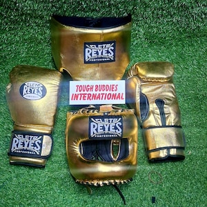 May include: A set of gold boxing equipment, including gloves, headgear, and a protective cup, all with the "CLETO REYES" logo. The items are arranged on a green surface.