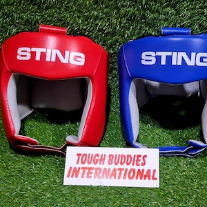 Op de afbeelding: Twee bokshelmen, &eacute;&eacute;n rood en &eacute;&eacute;n blauw, met het witte woord "STING". Ze liggen op een groene ondergrond, met een wit bord met de tekst "TOUGH BUDDIES INTERNATIONAL".