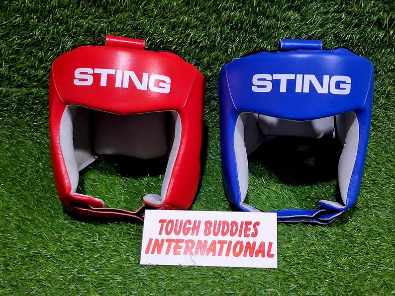 Op de afbeelding: Twee bokshelmen, &eacute;&eacute;n rood en &eacute;&eacute;n blauw, met het witte woord "STING". Ze liggen op een groene ondergrond, met een wit bord met de tekst "TOUGH BUDDIES INTERNATIONAL".