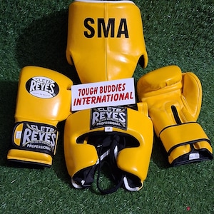Peut inclure: Ensemble d'équipement de boxe jaune, comprenant des gants, un casque et une protection thoracique. Le casque porte l'inscription "REYES PROFESSIONAL" et "M". La protection thoracique porte l'inscription "SMA". Les gants portent l'inscription "REYES" et "16 oz".