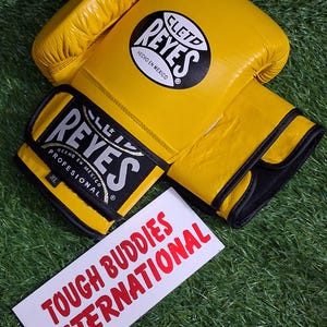 May include: Yellow boxing gloves with black accents and the brand name "REYES" in white and black. The gloves are on a green surface, with a white sign that reads "TOUGH BUDDIES INTERNATIONAL" in red.