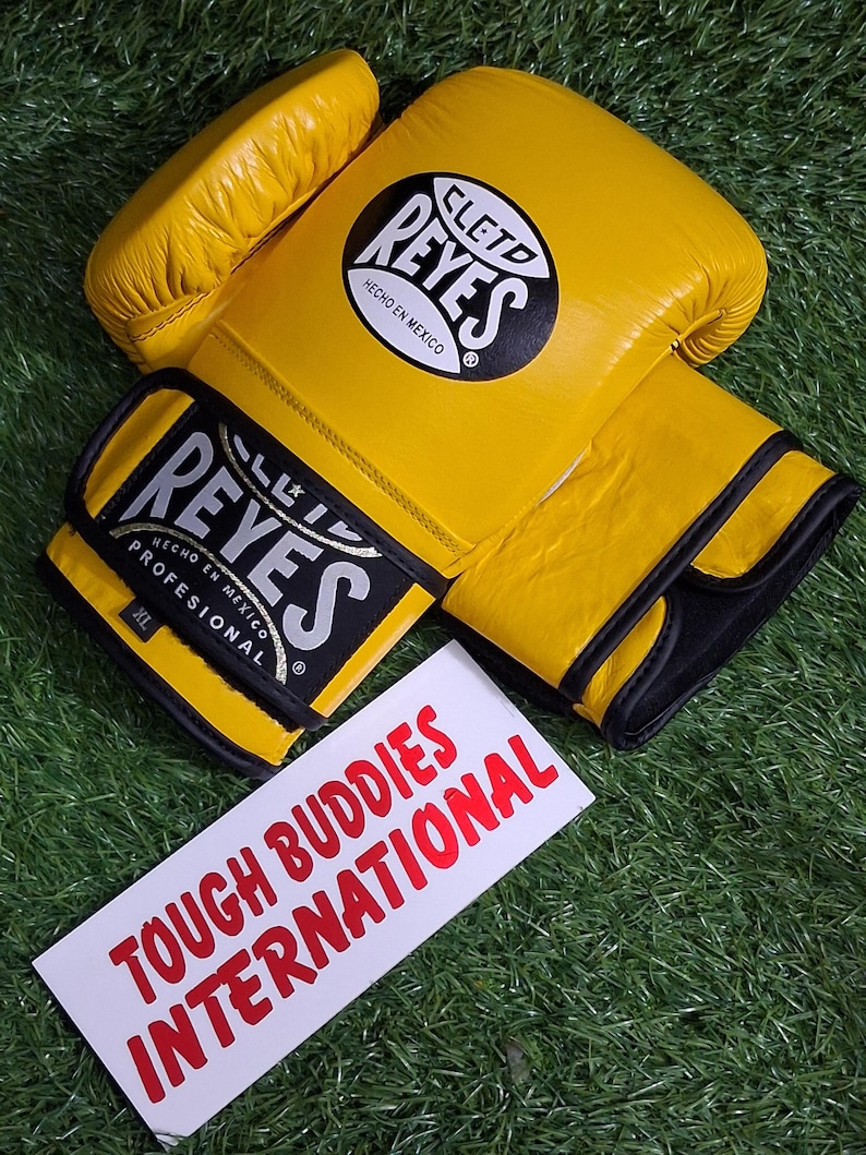 May include: Yellow boxing gloves with black accents and the brand name "REYES" in white and black. The gloves are on a green surface, with a white sign that reads "TOUGH BUDDIES INTERNATIONAL" in red.